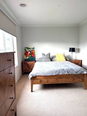 Quarto com cama de madeira, mesa de cabeceira com abajur, cômoda e um quadro colorido na parede.