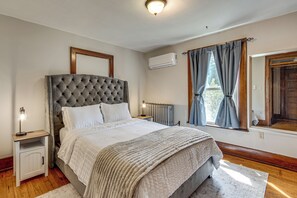 Una camera da letto con una testiera trapuntata, un letto con lenzuola bianche, un comodino con una lampada, una finestra con tende e un condizionatore.
