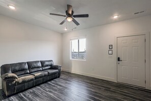 Une pièce avec un parquet en bois foncé, un canapé en cuir noir, un ventilateur de plafond, une porte blanche et une fenêtre avec des stores.
