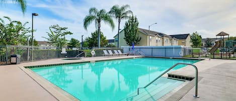 En swimmingpool med et skilt, der angiver en dybde på 107 centimeter, omgivet af et hegn, med en legeplads og huse i baggrunden.