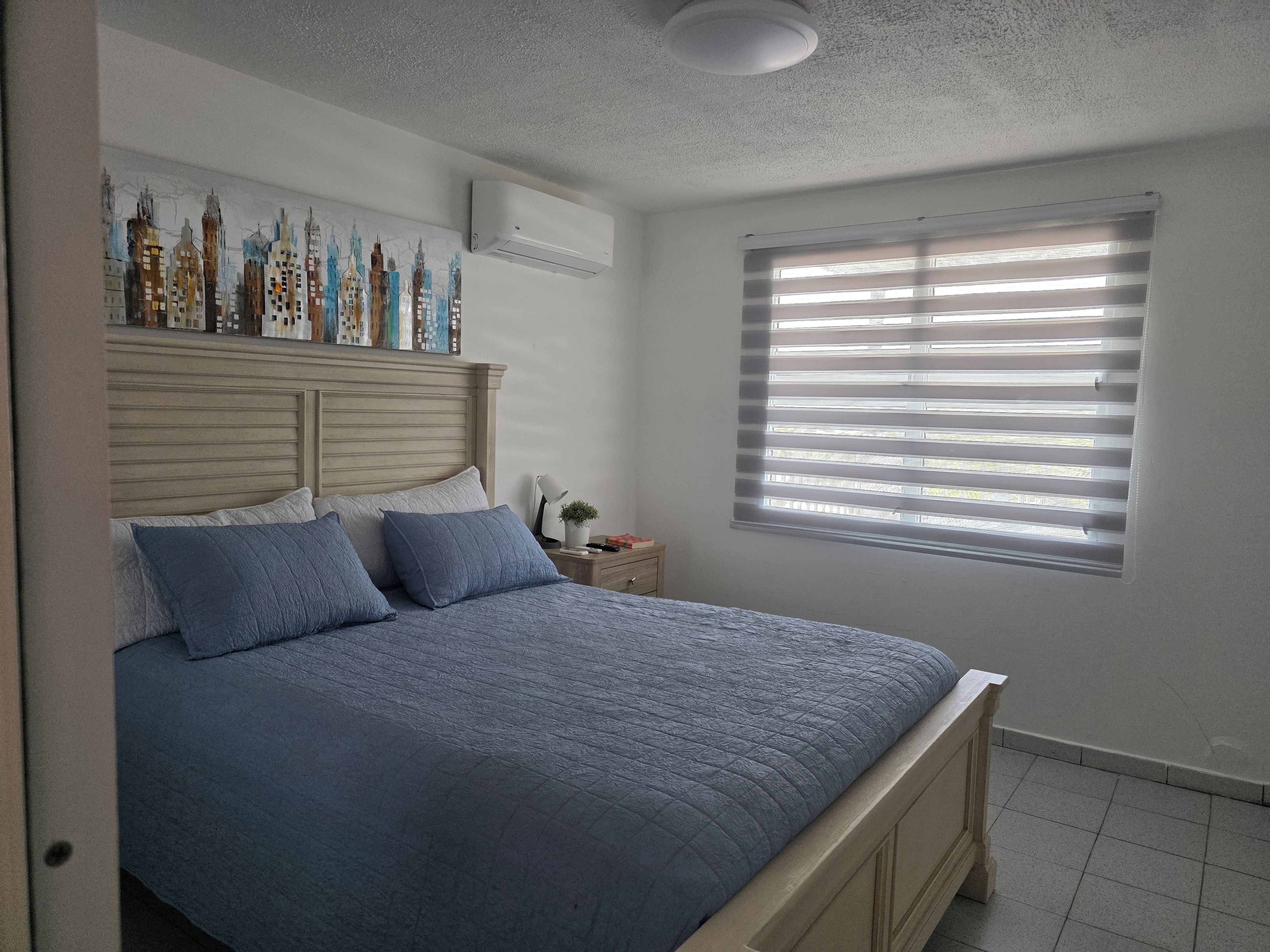 A bedroom with a bed, a nightstand, a wall-mounted air conditioner, and a window with blinds.