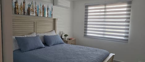 A bedroom with a bed, a nightstand, a wall-mounted air conditioner, and a window with blinds.