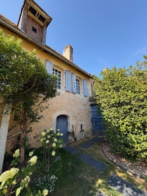 Ein zweigeschossiges Gebäude mit einer Sandsteinfassade, einem zentralen Eingang mit einer blauen Tür und einem kleinen Fenster mit Rollläden.
