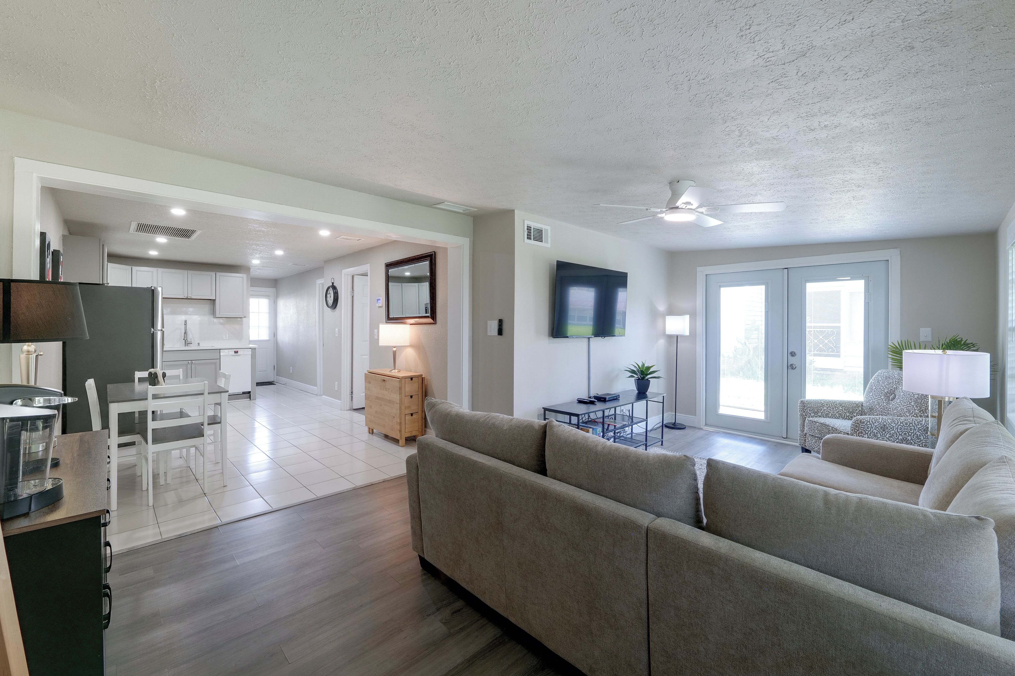 A modern living room with a grey sofa, a dining area with a table and chairs, a kitchen with white cabinets, and a large TV mounted on the wall.
