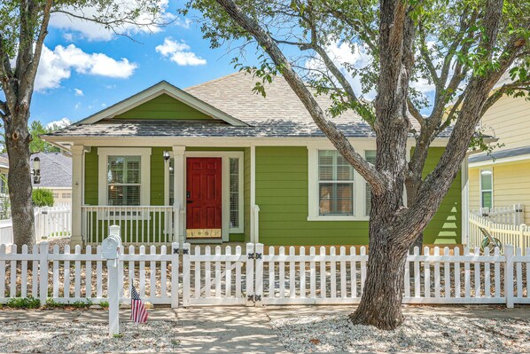 A small, single-story house with a green exterior, a red front door, and a white picket fence.