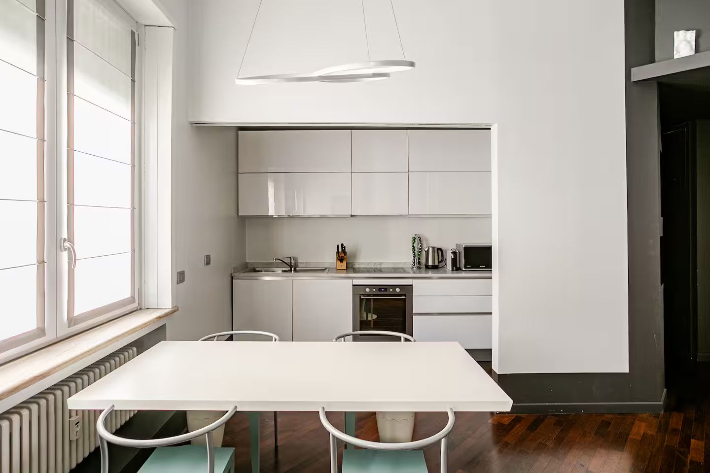 Una cocina moderna con armarios blancos, horno empotrado y un comedor con mesa y sillas blancas.