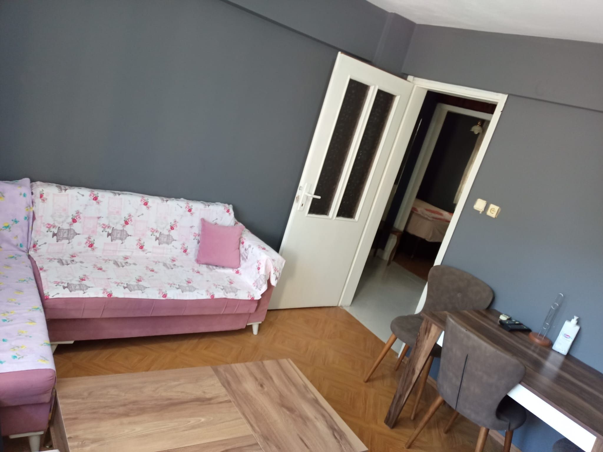 A room with a sofa, a wooden coffee table, a desk with a chair, and a door leading to another room.