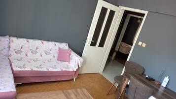 A room with a sofa, a wooden coffee table, a desk with a chair, and a door leading to another room.