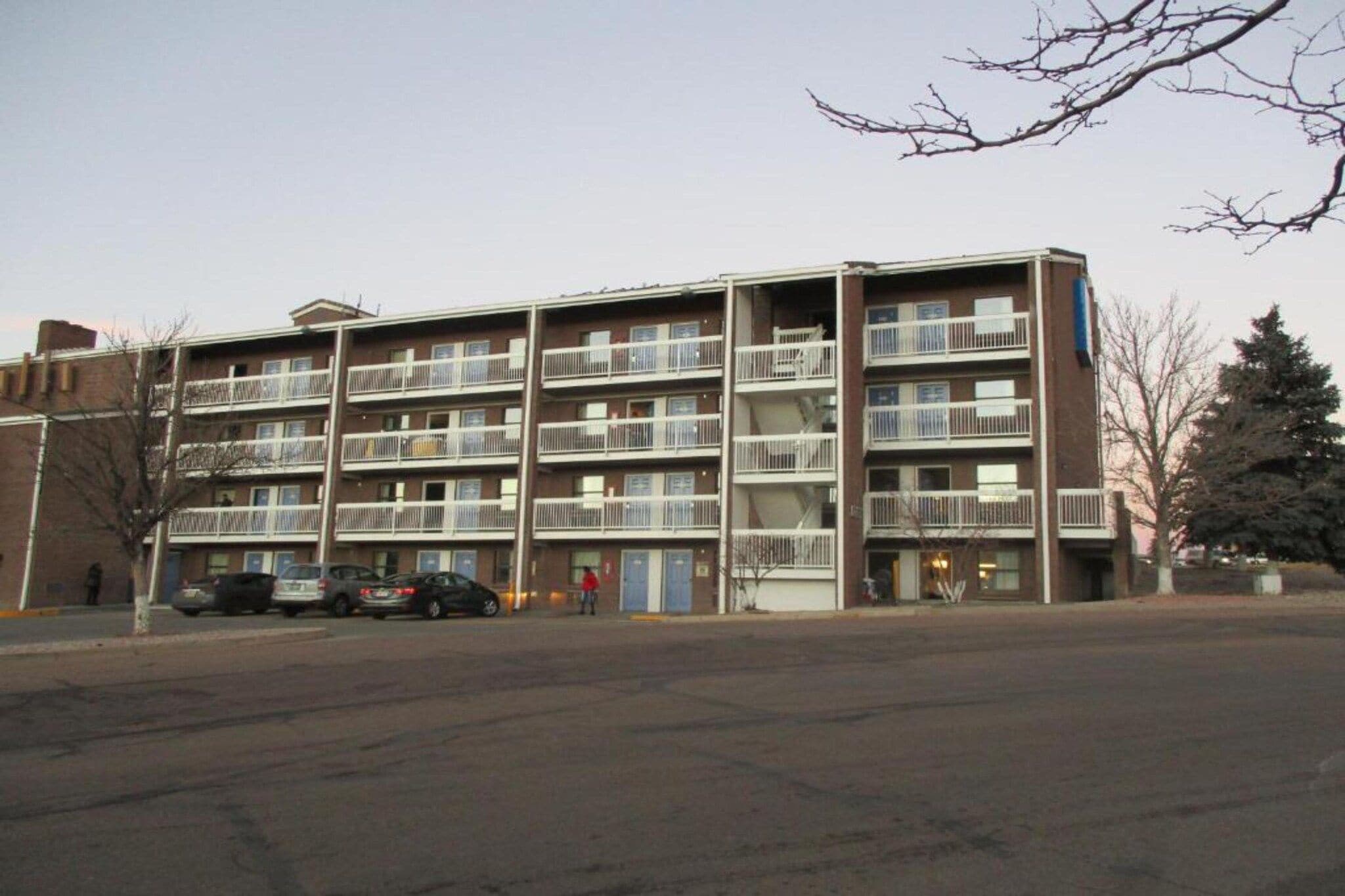 A multi-story apartment building with balconies, a parking lot, and a tree in the foreground.