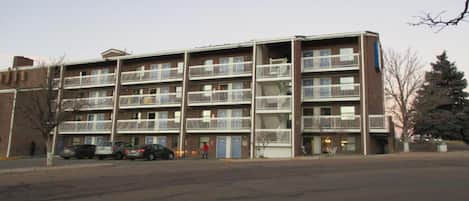 A multi-story apartment building with balconies, a parking lot, and a tree in the foreground.
