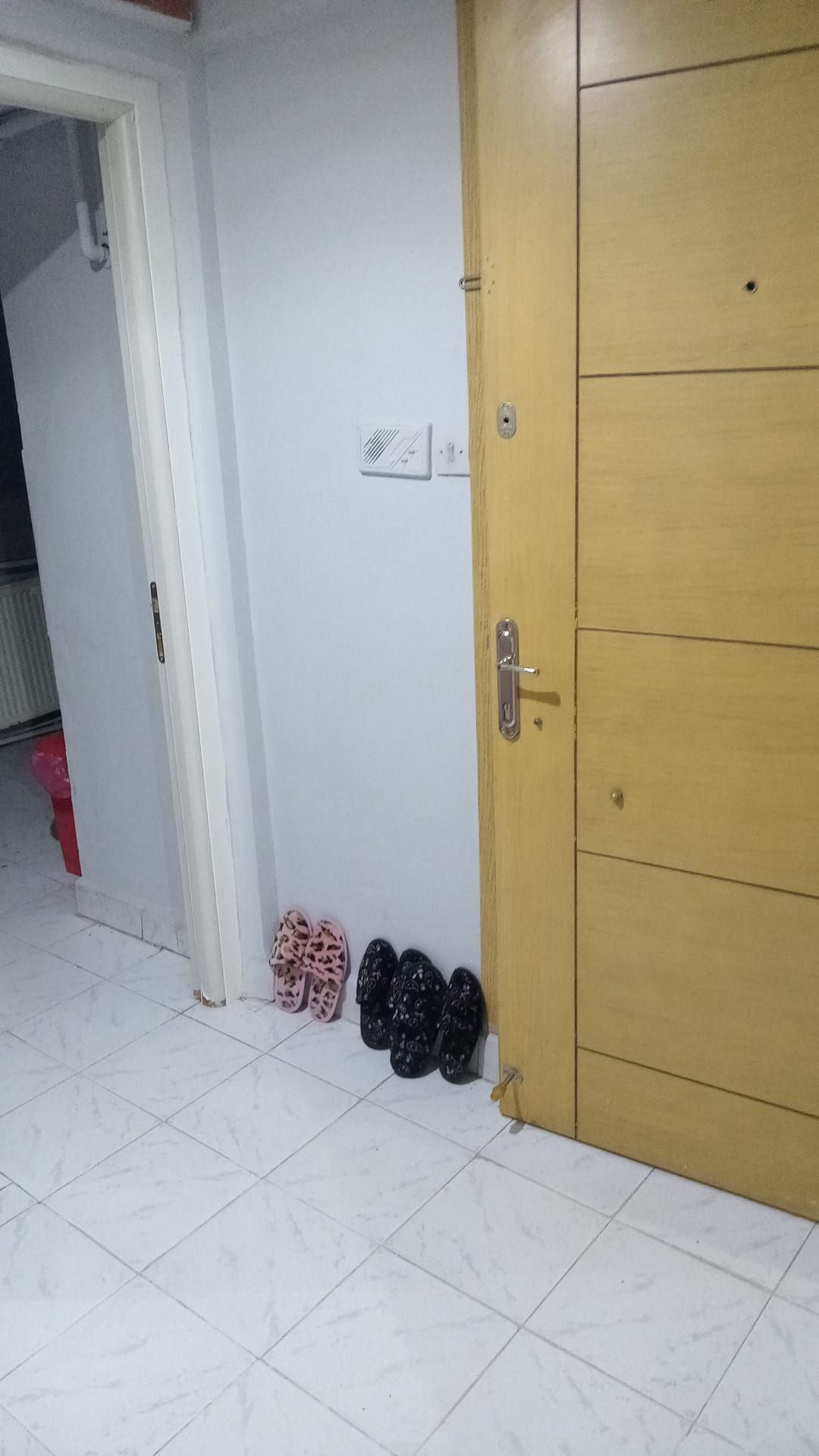 Une pièce avec un sol carrelé de carreaux blancs, une porte en bois et un mur blanc avec un interrupteur.