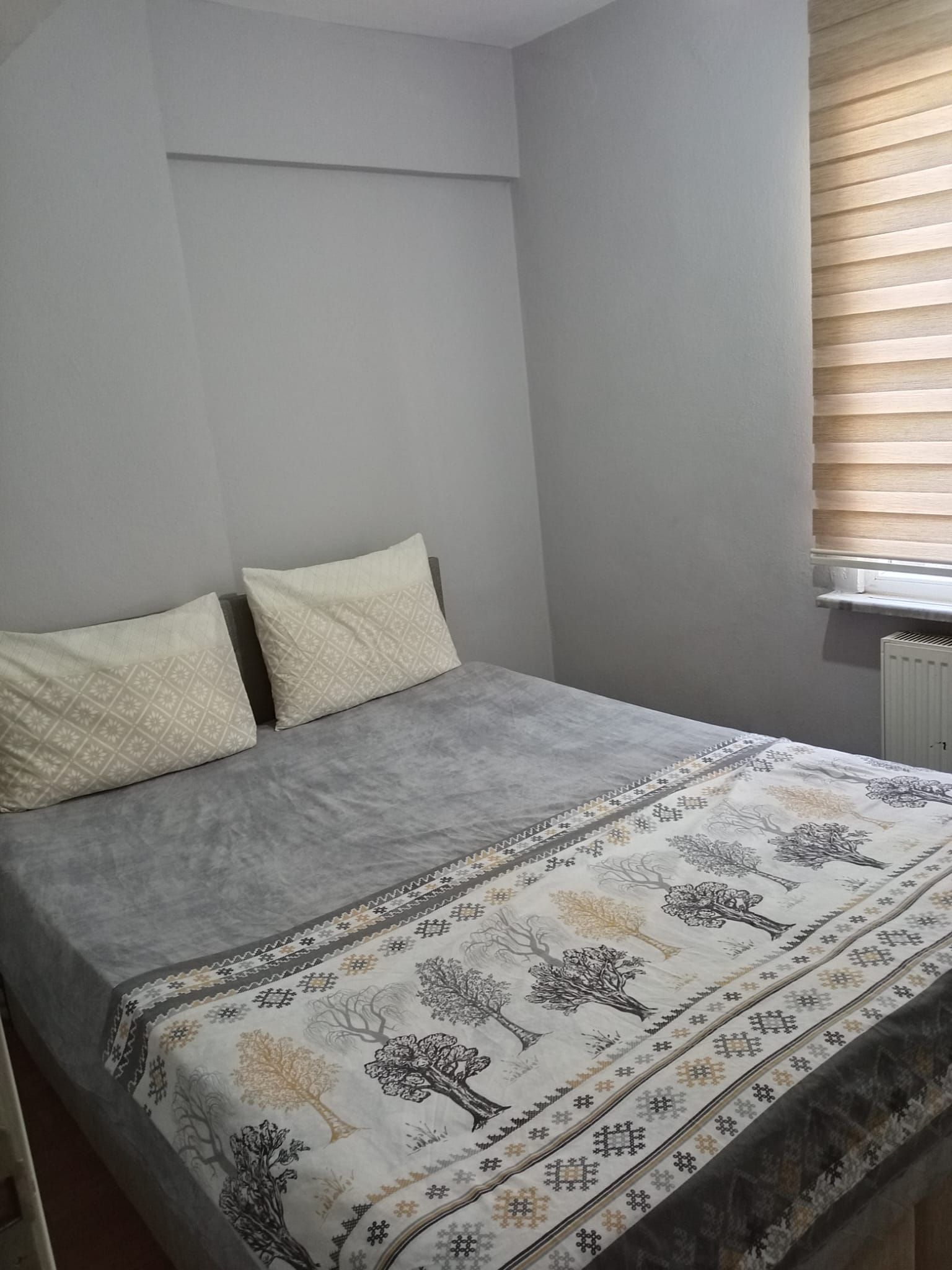 Un dormitorio con cama, dos almohadas, una ventana con persianas y un radiador.