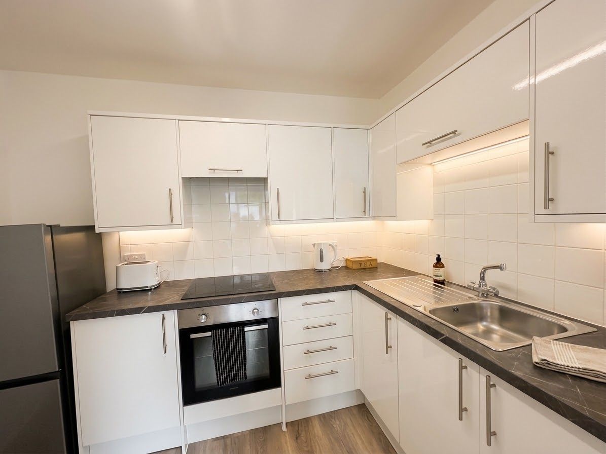 A modern kitchen with white cabinets, a black countertop, a built-in oven, a sink, and a kettle.