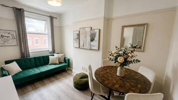 A living room with a green sofa, a dining table with chairs, a vase with flowers, and artwork on the walls.
