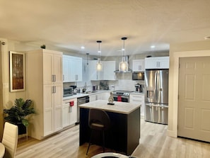 Una cocina moderna con armarios blancos, electrodomésticos de acero inoxidable, iluminación colgante y una isla central con asientos.