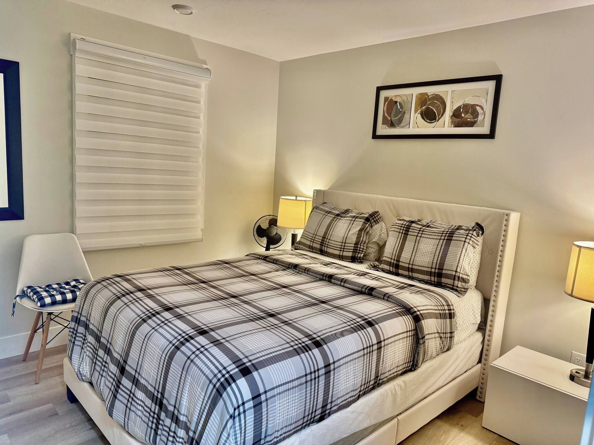 Una camera da letto con un letto, una sedia, un comodino, una lampada, un quadro appeso al muro e una finestra con persiane.