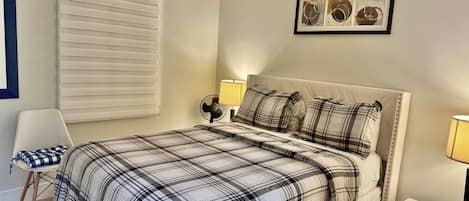Un dormitorio con cama, una silla, mesita de noche, lámpara, un cuadro en la pared y una ventana con persianas.