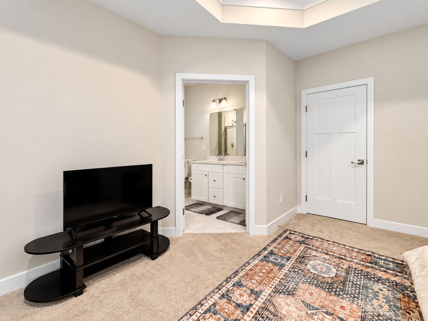 A bedroom with a TV, a patterned rug, a door, and a bathroom visible through an open doorway.