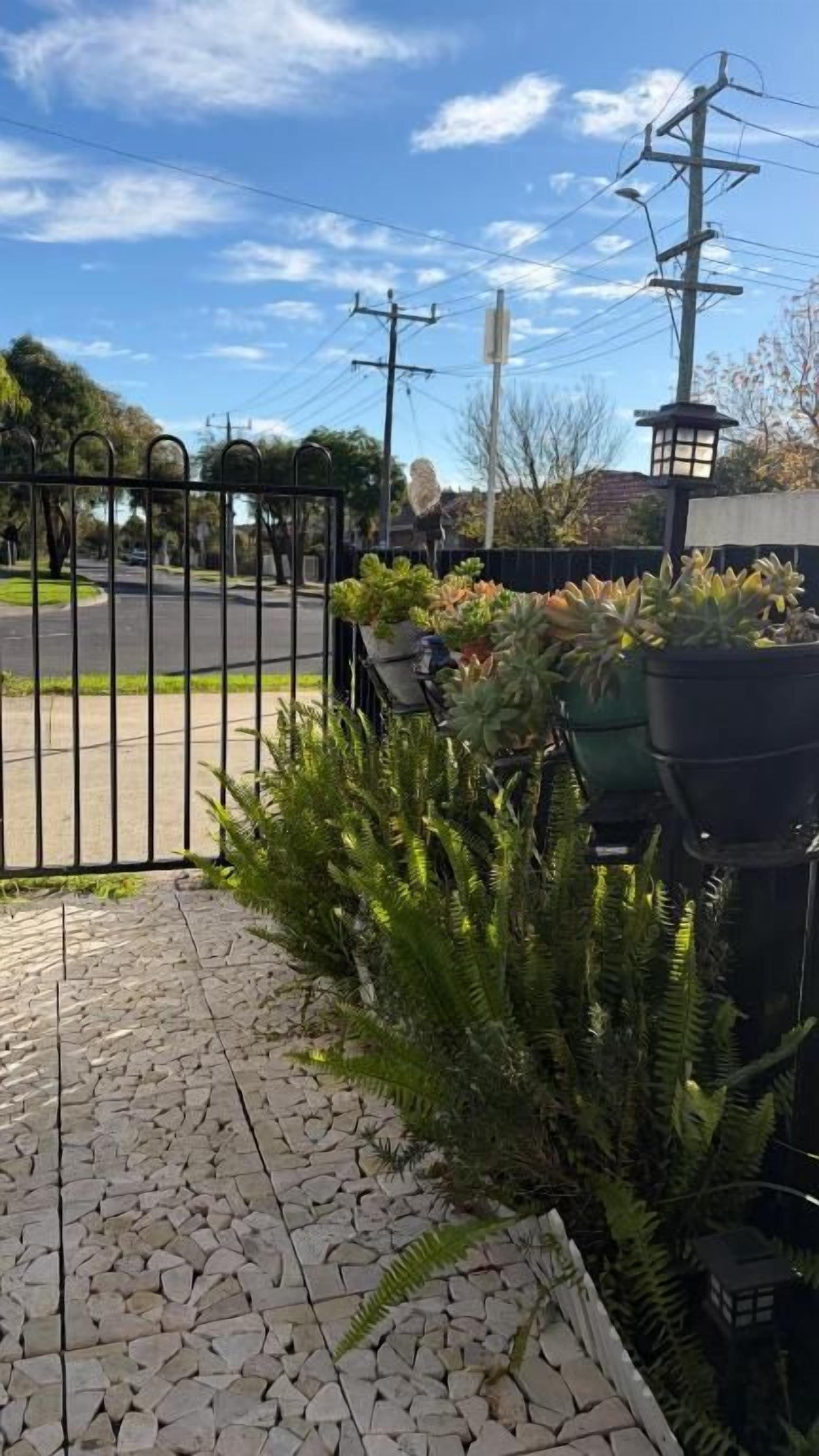Un chemin pavé mène à une entrée fermée par un portail, agrémentée de plantes en pot, avec une rue en arrière-plan.