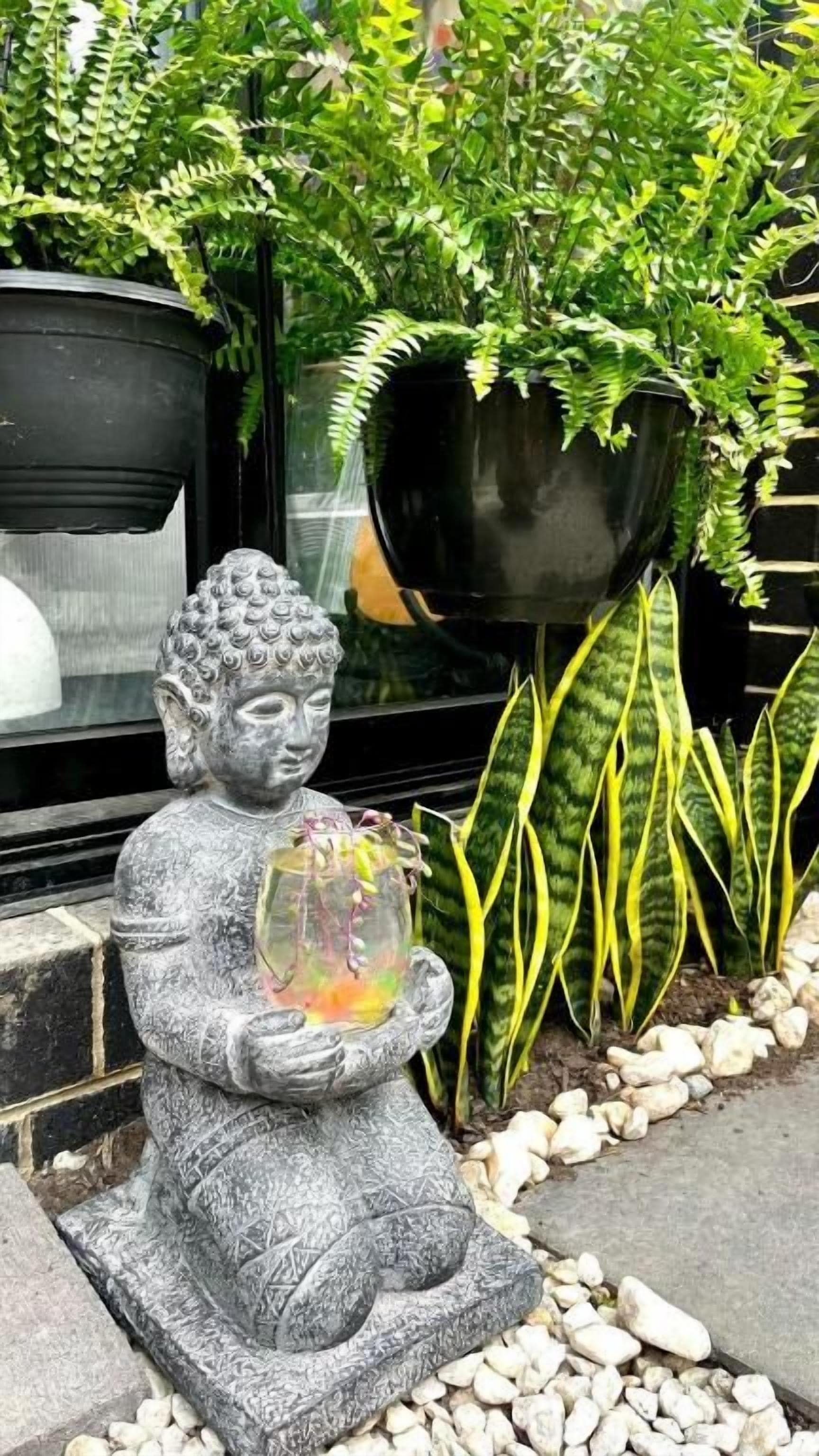A statue of a seated figure holding a fish bowl, surrounded by plants and a window.
