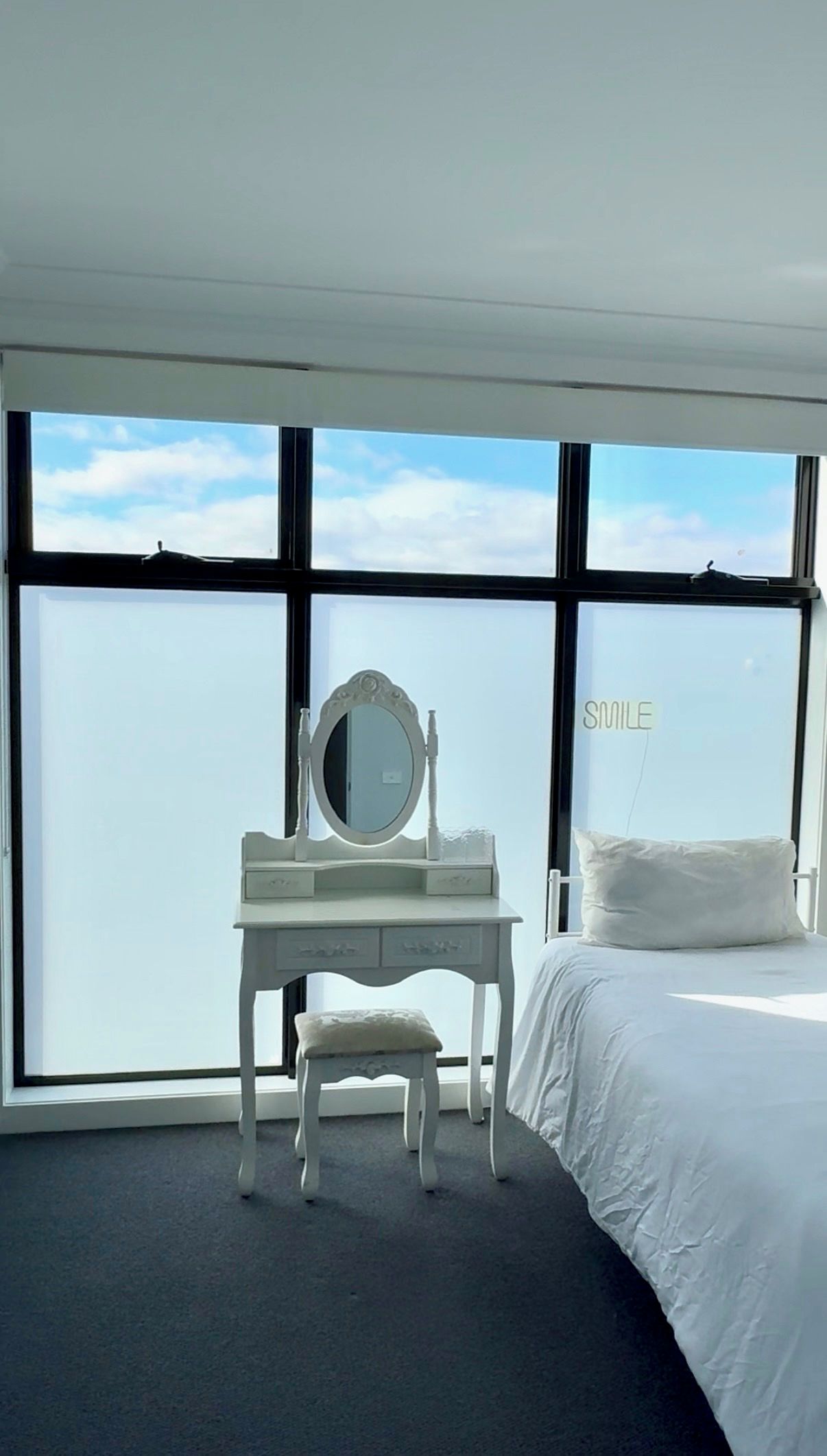 Une chambre d’hôtel avec un lit blanc, un bureau blanc doté d’un miroir et une grande fenêtre donnant sur le ciel.