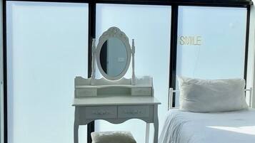Une chambre d’hôtel avec un lit blanc, un bureau blanc doté d’un miroir et une grande fenêtre donnant sur le ciel.
