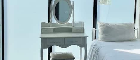 A hotel room with a white bed, a white desk with a mirror, and a large window offering a view of the sky.