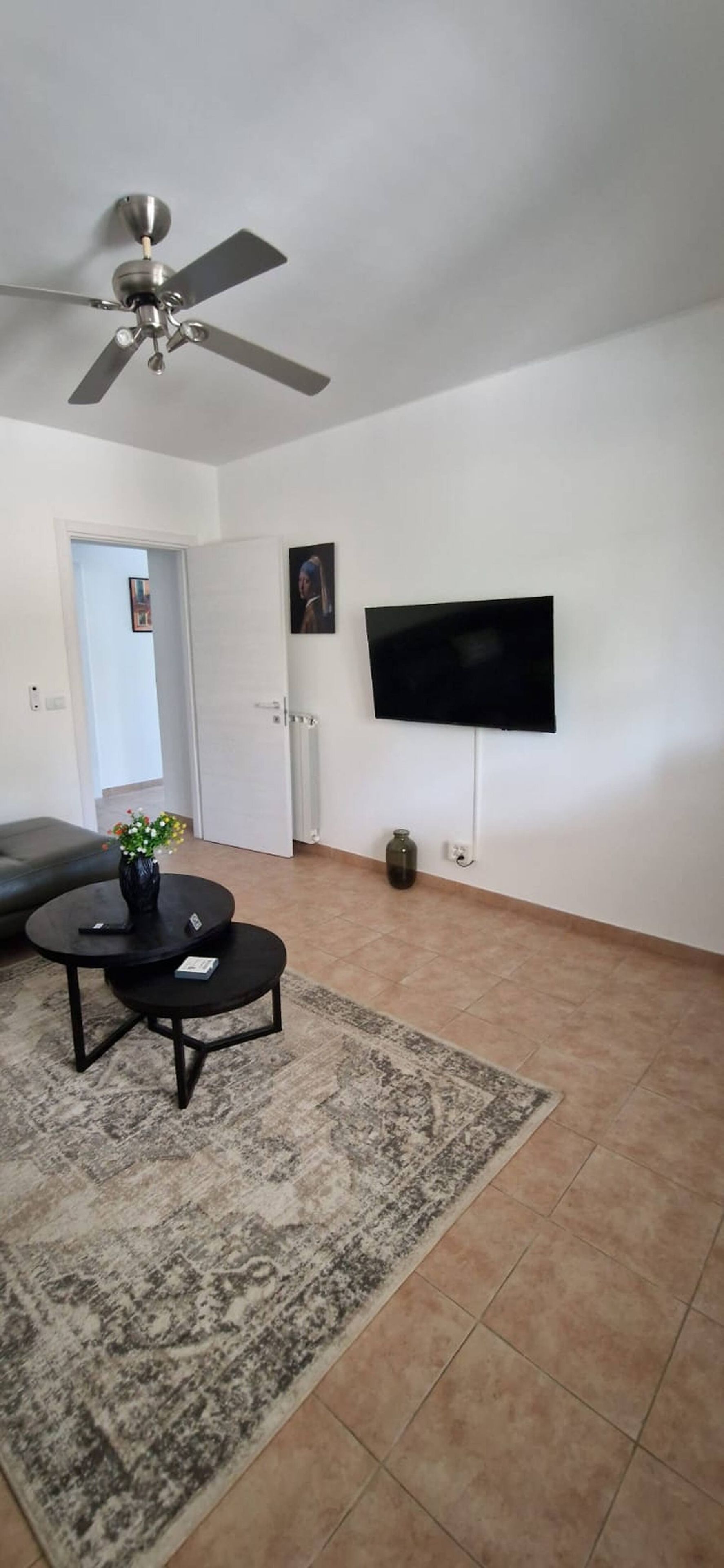A living room with a ceiling fan, a flat-screen TV mounted on the wall, a round coffee table with a vase of flowers, and a grey sofa.