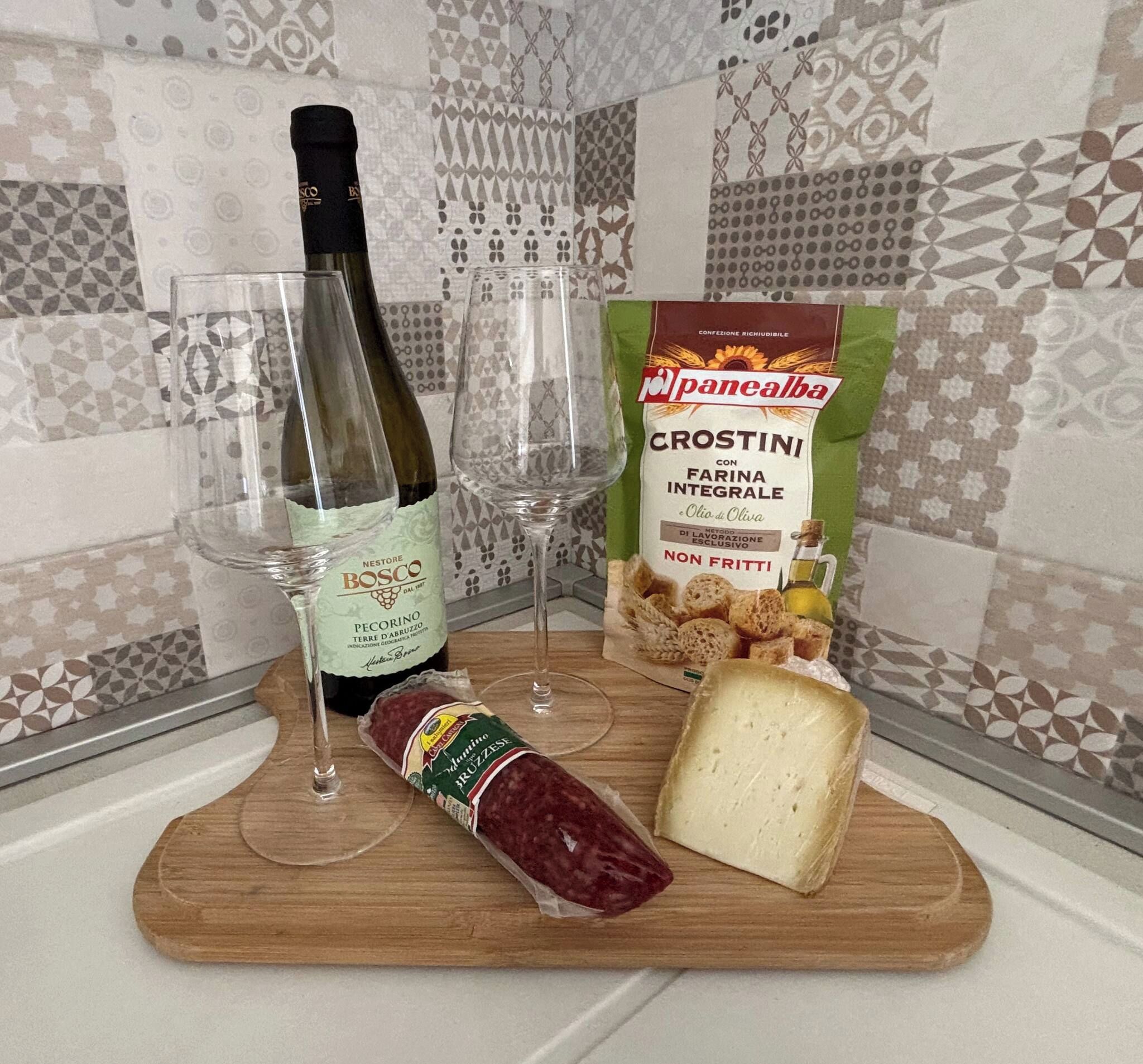 A wooden cutting board with a bottle of wine, a glass, a package of bread, and a block of cheese.