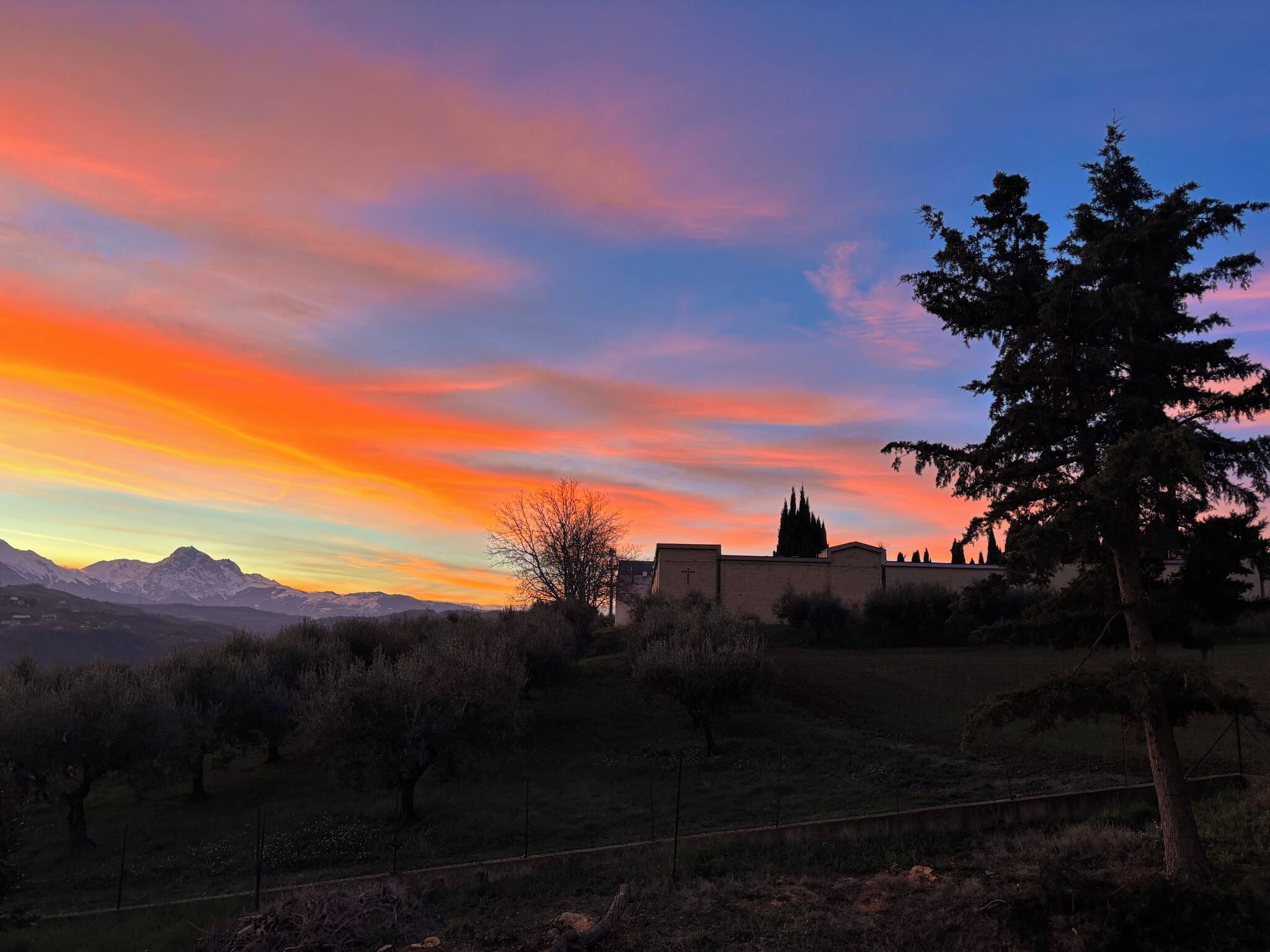 Un coucher de soleil sur un paysage montagneux, avec une église et un arbre au premier plan.