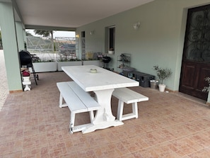 Un patio con mesa y banco blancos, una parrilla y plantas en macetas.