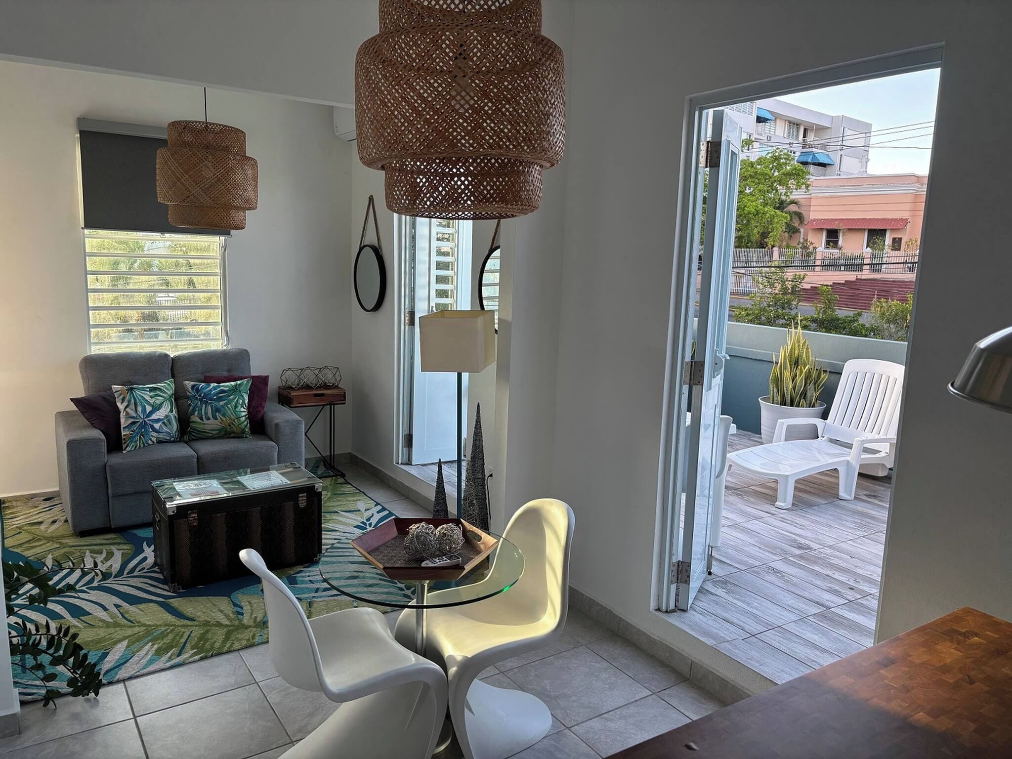A modern living room with a sofa, coffee table, and chairs, a balcony with outdoor furniture, and a large window providing a view of the city.