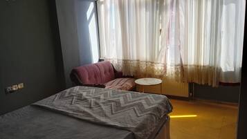 A bedroom with a bed, a chair, a small table, and a window with sheer curtains.