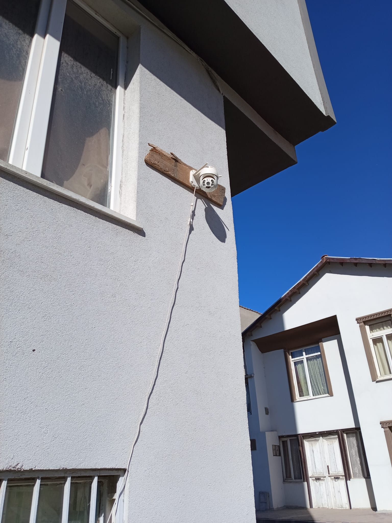 Un racó d'edifici amb façana blanca, una finestra i una càmera de seguretat muntada sobre un biga de fusta.