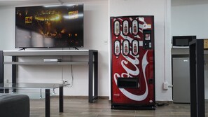 A room with a television mounted on the wall, a Coca-Cola vending machine, a table with a glass top, and a refrigerator. - Modern Loft King Bed, Wifi & Smart Lock (Mexico City)