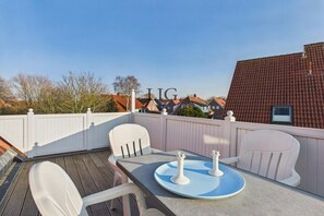 A deck with a table and two chairs, a white fence, and a building with a red roof in the background. - Holiday apartment Uhlenkamp 7 (Langeoog)