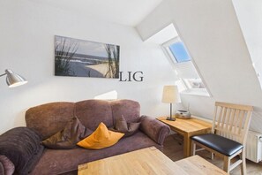 A room with a brown sofa, a wooden table, a chair, a lamp, and a picture on the wall. - Holiday apartment Uhlenkamp 7 (Langeoog)