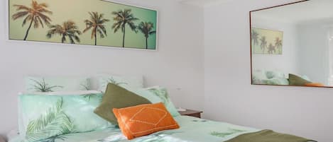 A bedroom with a bed, pillows, a mirror, and a painting of palm trees.