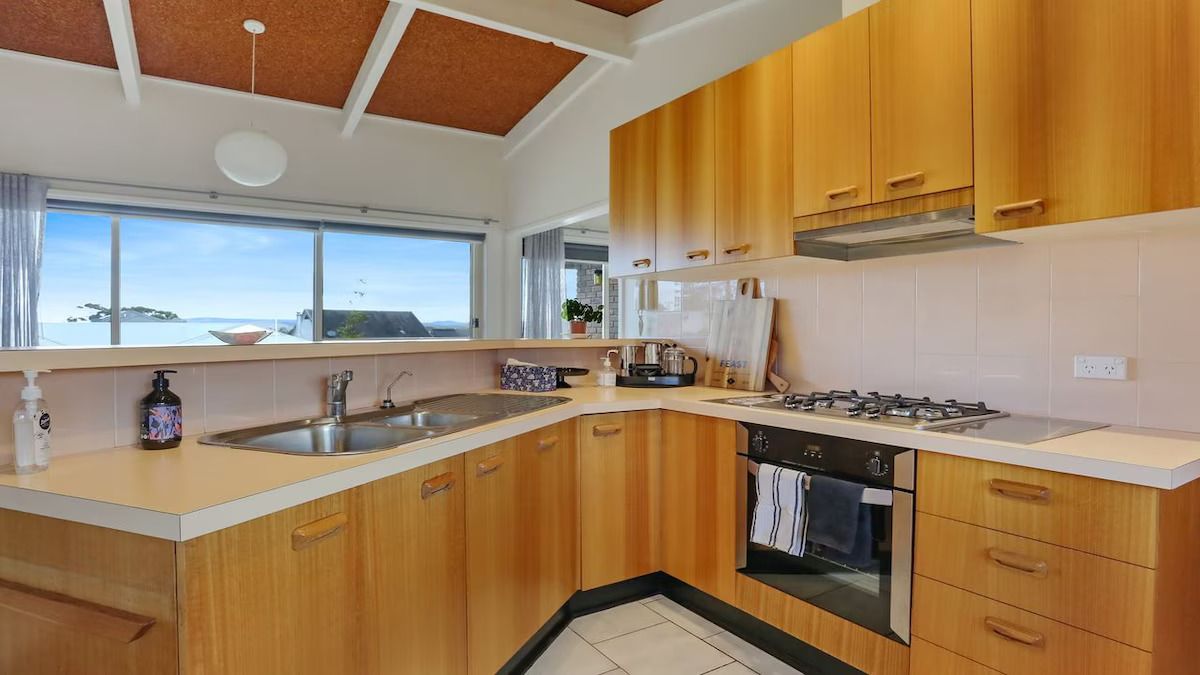Cocina moderna con armarios de madera, estufa a gas, pileta y vista al mar a través de amplias ventanas.