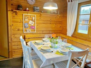A wooden cabin interior with a dining table set for four, white chairs, a clock on the wall, and a window with curtains. - Holiday Home Neckar 58 (Hayingen)