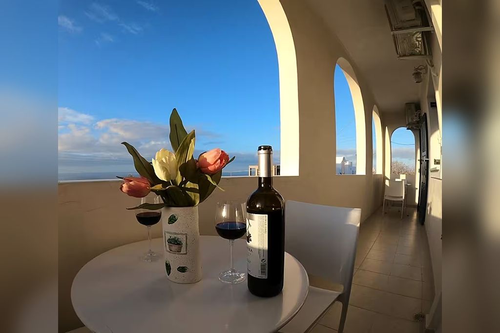 A balcony with a table set for two, a bottle of wine, and a vase with flowers.