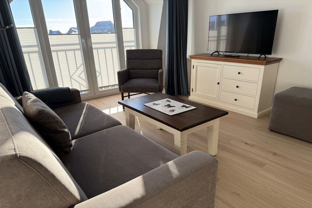 Uma sala de estar moderna com sofá, poltrona e mesa de centro. Uma TV de tela plana está fixada na parede acima de um armário.