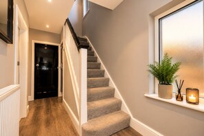 A staircase with carpeted steps leading to a landing, a black door, and a window with a plant and candle on the sill. - Modern 3-Bed, 2-Bath Home | Parking for 3 | Air Conditioning | Rugby (Rugby)