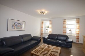 A living room with two black leather sofas, a wooden coffee table, a ceiling fan, and a framed floral artwork on the wall. - Holiday apartment Greetsiel (Osteel)