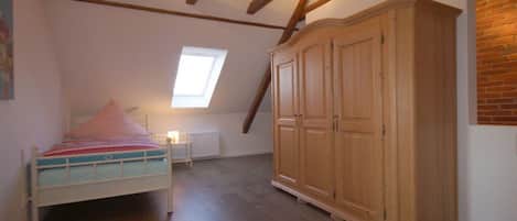 Ein Schlafzimmer mit Bett, Schrank, Heizkörper und einer geneigten Decke mit freiliegenden Balken.