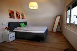 A bedroom with a bed, bedside table, lamp, artwork, and a window. - Holiday home with 3 bedrooms (Heiligenhafen)