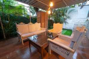 An outdoor seating area with a wooden table and white cushioned chairs, surrounded by lush greenery and a stone wall. - The signature crest (Wai)