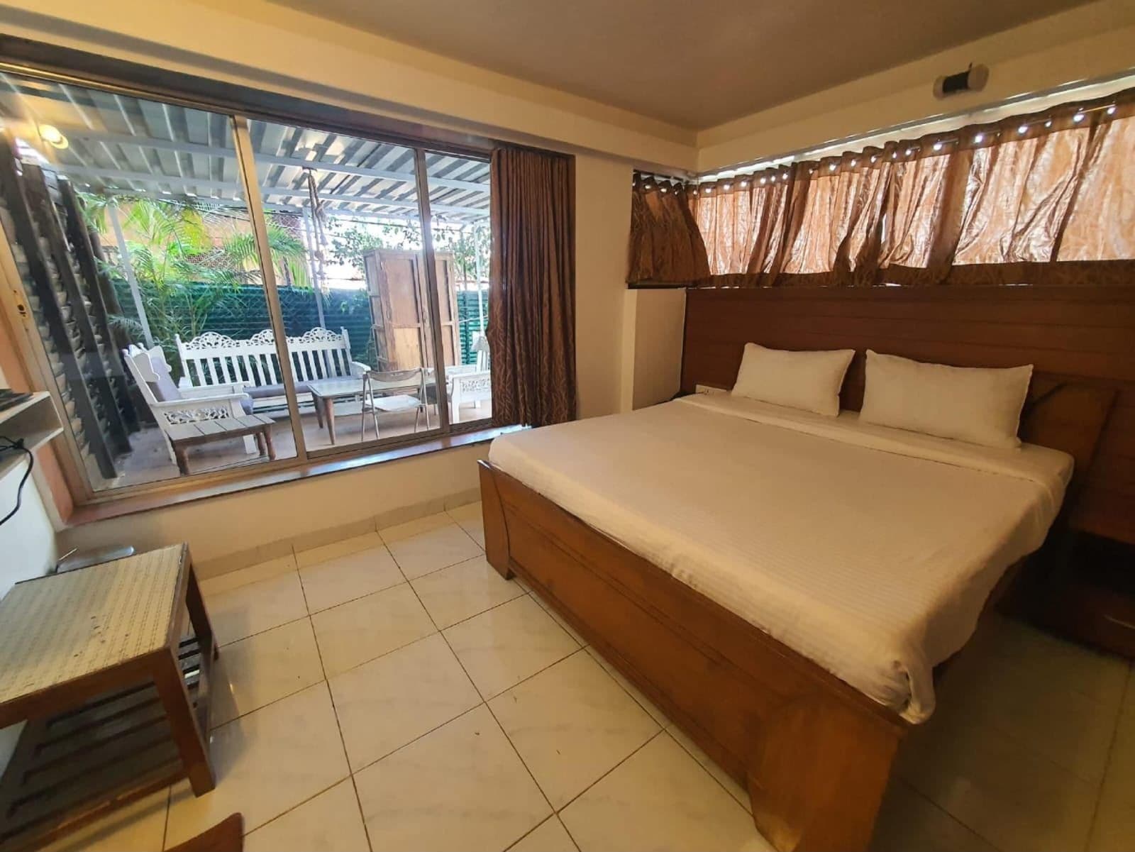 A bedroom with a large bed, a window with curtains, a small table, and a view of an outdoor patio with chairs and a white fence.