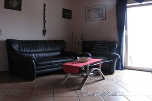 A living room with a black leather sofa, a glass coffee table with a red top, and a black chair. - Holiday apartment with 1 bedroom (Weibern)