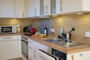 A modern kitchen with white cabinets, a wooden countertop, a stainless steel sink, a microwave, a red kettle, a bottle of wine, and a dish soap dispenser. - MS Apartments | Sielterrasse 1 (Ditzum)